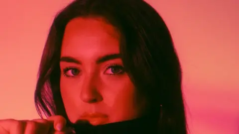 Ellie Scougall has long dark hair and is holding a dark-coloured top over her chin but below her mouth. The image has a red filter on it 