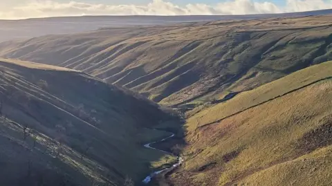 A green valley with a river winding through its centre and hills in the background against a clouded skyline.