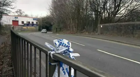 BBC A metal fence beside a road with blue and white police tape knotted around it and trees on the other side