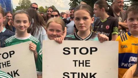 Four children are standing in a line holding placards saying stop the stink. They are also wearing football jerseys.