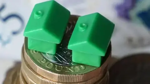 Two small plastic green house models sit on top of a stack of pound coins, there is a £10 and a £5 note in the background.