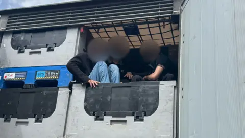 Image of three men sat on grey and black metal boxes looking out the back of a lorry.