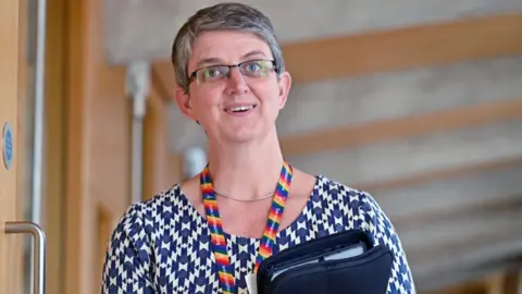 Getty Images Maggie Chapman, who has short grey hair and glasses, looks towards the camera while holding a folder to her chest. She is wearing a patterned blue and white top, with a rainbow-coloured lanyard. 