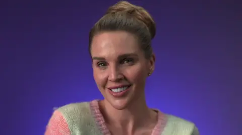 Danielle Lloyd smiles wearing a multicoloured cardigan; she has brown hair tied in a bun