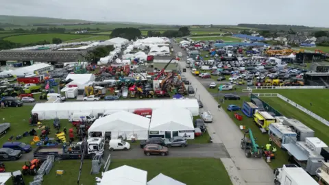 An aerial view shows a large outdoor agricultural show with rows of white tents, machinery displays and parked vehicles spread across a field.
Crowds, tractors, food vans and exhibition stands are arranged along wide pathways, with farmland and rolling countryside stretching into the distance.