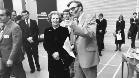 The Wood Family A black and white image of Sir Ian Wood, in light suit, showing Margaret Thatcher around a hall. He is pointing and she is smiling