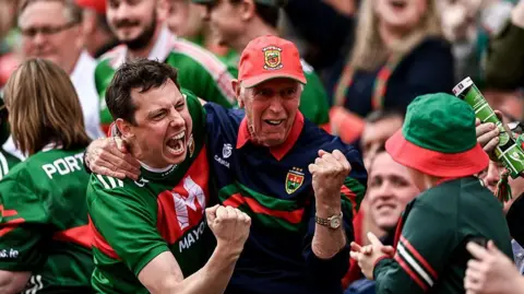 Getty Images A crowd of people. In focus are two men with their arms around each others' shoulders. They are both wearing red and green sports tops. 