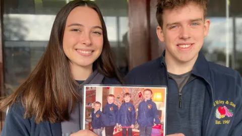 Simon Thake/BBC A young woman with long brown hair smiles while holding up a picture. The boy next to her also smiles. Both wear blue fleeces.