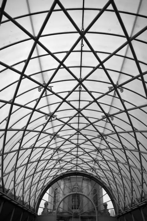 SARAH CALLOW A black and white picture of a curved roof with criss-cross metal bars across it creating a tunnel that leads to a large building at the far end.