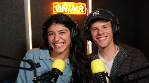 Movers+Shakers Lilia Souri and AJ Pulvirenti who co-host a Gen Z marketing podcast. Smiling they are setting in a studio in front of microphones.