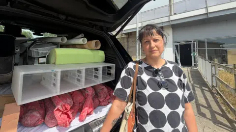 BBC/Jim Scott Joanna Manousis, an artist from Ohio, wears a white top with black circles on it and is holding a brown bag. She has just cleared equipment from her workshop inside the National Glass Centre and is standing in front of the boot of a car, which has been opened and now contains a plastic shelf which stored her tools, and multiple red cylindrical bags of materials.