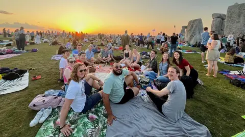 CircleUp A group of people sitting on blankets under a sunrise at Stonehenge. They are all looking at the camera and smiling. Large crowds can be seen in the distance while Stonehenge is to the right of the frame.