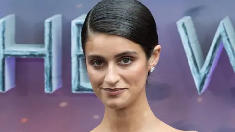 Getty Images A woman with slicked back dark hair, parted to the side, stands in front of a hoarding on a red carpet. She's wearing expensive-looking diamond stud earrings.