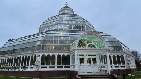 A close-up shot of the Palm House, a domed glass conservatory surrounded by parkland in Sefton Park.