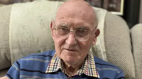 Don Way holds a cup off coffee in his right hand while sitting on his cream coloured sofa. The cup has a picture of a red squirrel on it 