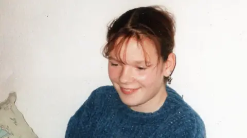 A picture of Lindsay Rimer as a child. She is sat down smiling with her brown hair tied back, wearing a blue jumper.