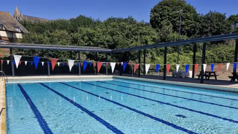A view of a pool. The pool water is a clear blue and is very still. There are dark lines at the bottom of the pool mapping out the lanes. Red, blue and white bunting hangs above the pool. Trees surround the outside of the pool site.