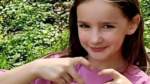 Family photograph A girl with long brown hair and a pink top makes a heart symbol with her hands.