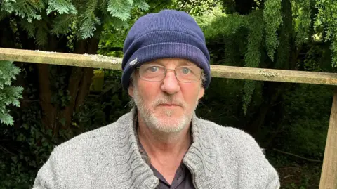 Man in a blue beanie wearing glasses with a beard and moustache with green foliage around 