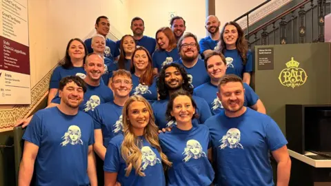A group of 20 people stand in a theatre corridor. They are wearing blue T shirts with white logos on. They are smiling at the camera.