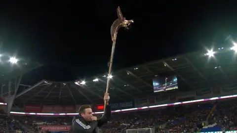 S4C A man stands on a football pitch playing a long, bronze horn.