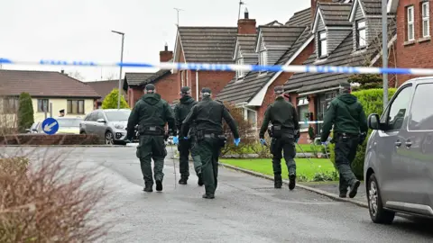 Pacemaker Five police officers wear green and black uniforms with black hats. They are walking up a concrete residential street with red brick houses on their right hand side, they have their backs to the camera. 