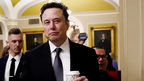 Reuters Elon Musk walks through Capitol Hill holding a takeaway coffee in his hand and looking away from camera, wearing a suit and tie and followed by security officers and journalists, in Washington DC on 5 December.