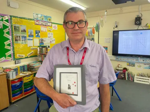 Gemma Dawson/BBC Headteacher Charlie Hebborn. He has short grey hair and is wearing a purple shirt. He is standing in a classroom holding a card set in a picture frame.