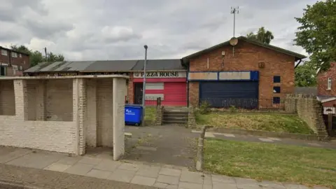 A single storey row of shops which look run down and one is painted red with a black and white sign readings 'pizza house'. There is a rick bus shelter in the foreground. 