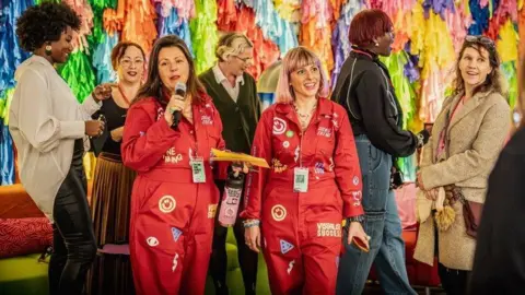 Two people waring red boiler suits covered in patches and images. The person on the left is speaking into a microphone. They are surrounded by a group of people and stood in front of a colourful, textile backdrop.