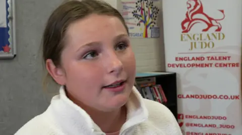 Stevie, a young girl in a white fleece top is speaking and standing against a wall with a bookcase and a sign which reads "England Judo" and says underneath that this is an England talent development centre.