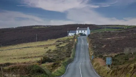 The Mountain Road stretching uphill up towards Kate's Cottage, which is a white cottage on the left, from the Creg ny Baa. Heathland can be see on both sides.