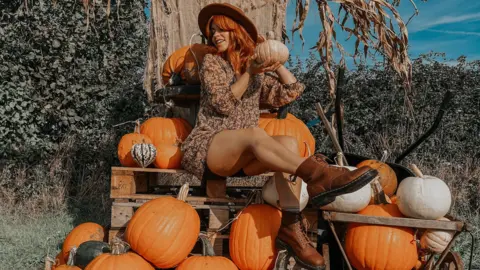 Picking Patch A woman with red hair sits on top of a wooden crate, surrounded by pumpkins. She is wearing a hat, red boots and a dark patterned sun dress