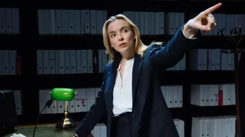 Jodie Comer pictured standing at a desk and pointing whileacting in Prima Facie