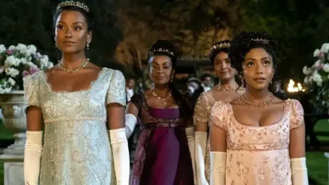 Netflix A still from Bridgerton shows Simone Ashley as Kate Sharma, in a beautiful pale green Regency beaded gown and ivory evening gloves, with her sister in the show, Charithra Chandran playing Edwina Sharma, in a similar-style gown in a pale pink. Adjoa Andoh as Lady Danbury is behind the two in a deep purple gown.