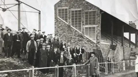 A black and white image of fans standing next to the stand at Borough Park in 1937, The stand is a brick construction with steps at the front leading up to the seats. The fans are standing on a mud bank. There are a few boys near the front while the rest are men all wearing hats.