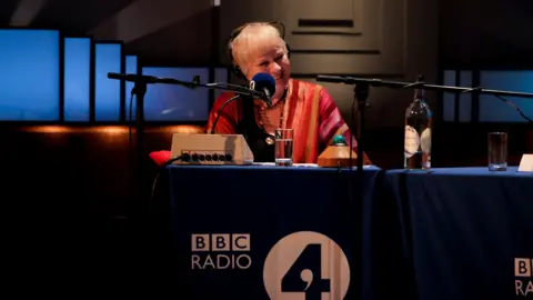 Tricia Yourkevich/BBC A woman sits at a table with a microphone and a large BBC Radio Four logo on the front.