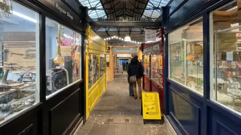A view of the inside of St Nicks Market. The image shows a narrow indoor corridor with a paved floor. On each side are shop windows and coloured shop fronts. Two people are looking in windows.