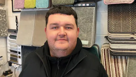 Vaughn Corrie, 27, is pictured in his carpet shop at the Civic Centre in Wythenshawe.