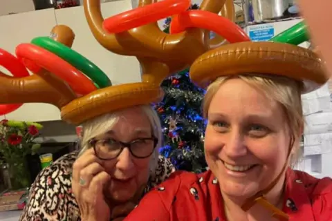 Clare Jones Photograph of Clare Jones (right) and her mum (left). Clare smiles at the camera, has blonde hair and blue eyes. Her mum wears black square rimmed glasses and also smiles at the camera, holding her left hand up to her eyes. They both wear inflated reindeer ears and a christmas tree can be seen behind them. 