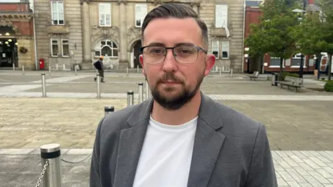 Connor Naismith - a man with short dark hair, glasses, a beard, grey jacket and white shirt - stands in a town square.