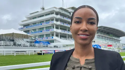 Julia Gregory/ BBC A woman wearing a black blazer and grey top. She is stood in front of a racecourse grandstand.