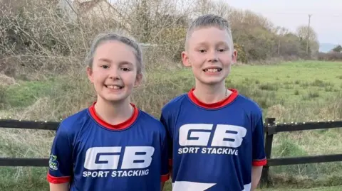 Two children in sports uniforms smiling with their hands behind their backs while outside in front of a field.