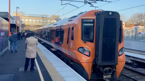 An orange and purple train on a platform. We can see a Willenhall sign in the background. It is a bright day and the sky is a clear blue. There are lots of people on the platform. We can see one man in a beige overcoat and a hat. 