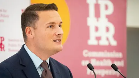 A side profile of PA Media Health Secretary Wes Streeting was wearing a Navy suit, blue shirt and brown tie in a microphone in a podium. She is clean with small, black hair.