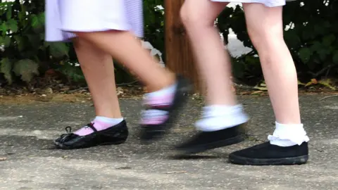 A blurred photo of schoolgirls walking to school. The photo just shows their skirts, legs and black school shoes