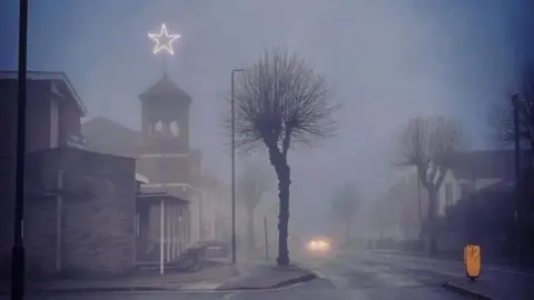 A church on a misty street in the evening. There is an illuminated white star on its tower. There are trees and residential buildings on the street.