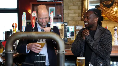Getty Images Prince William concentrates as he pulls a pint from behind the bar at a pub as another man, who is smiling, looks on