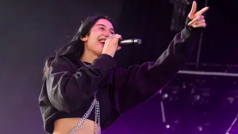 Julia Wolf performs on stage. Dressed in black, with metal chains, she is smiling while singing into a microphone