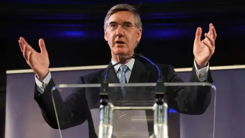 PA Media Jacob Rees Mogg standing in front of a glass podium, opening his arms and hands as he addresses an audience through a microphone. He is wearing a black jacket, white shirt and a silvery-coloured tie